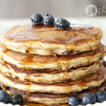 A stack of oat pancakes sit on a plate with blueberries on top